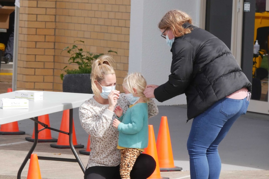 A blonde-haired woman kneels in front of a blonde child while another woman ties a face mask onto the child's face.