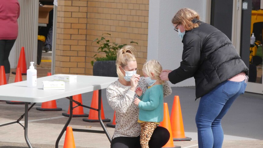 A blonde-haired woman kneels in front of a blonde child while another woman ties a face mask onto the child's face.