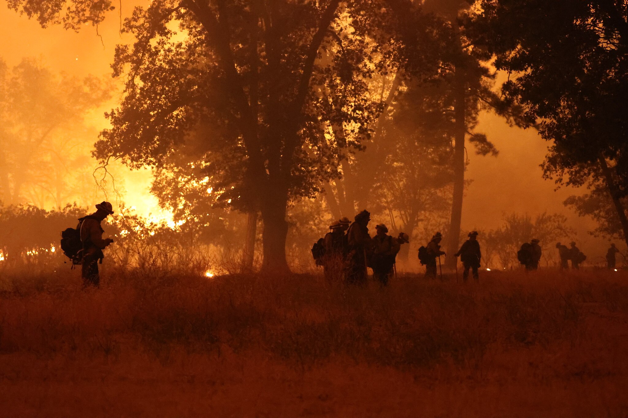 Firefighters walk through bushland in a haze of fire as flames burn in the background 