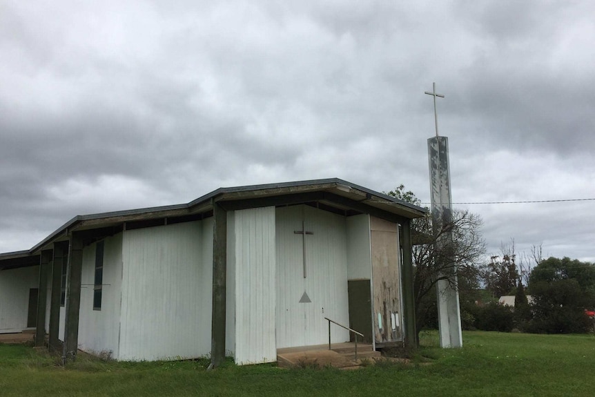 A white 1950s-style church on a green lawn with grey stormclouds in the sky.