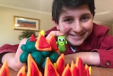 A smiling boy in a red shirt sits at desk behind tubs of Play-Doh and figures made of the substance.