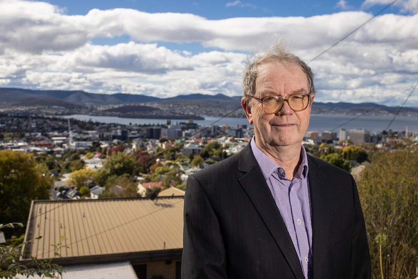 Un hombre con gafas y traje con la ciudad de Hobart al fondo.