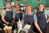 Staff and trainees stand behind the front counter at a coffee shop and smile at the camera.