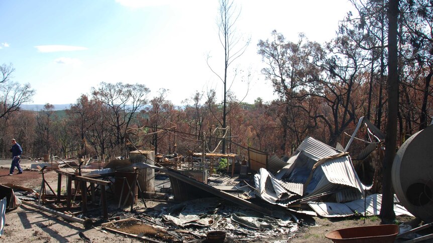 Sheets of metal lie on the ground next to a metal structure remains of a burnt out shed with charred forest in the background