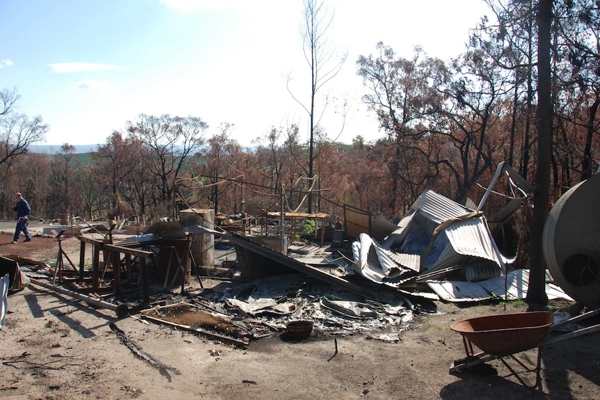 Sheets of metal lie on the ground next to a metal structure remains of a burnt out shed with charred forest in the background