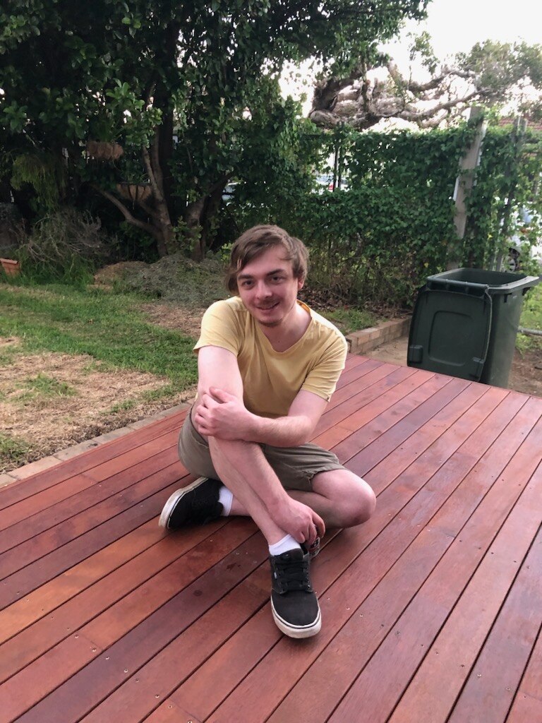 a young man sits on a wooden deck with his arms folded and one knee raised.