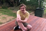 a young man sits on a wooden deck with his arms folded and one knee raised.