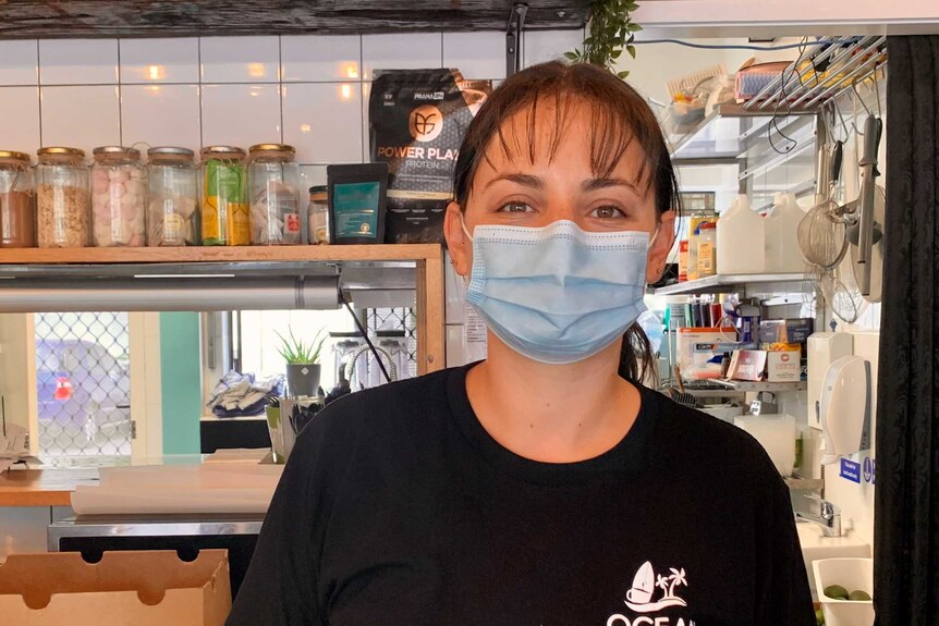 A woman in black t-shirt wears a mask standing in a cafe.