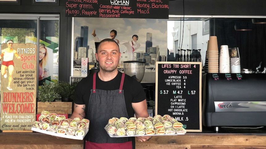 Steve Pace standing in front of the counter of his cafe holding two tray of sandwiches and wraps