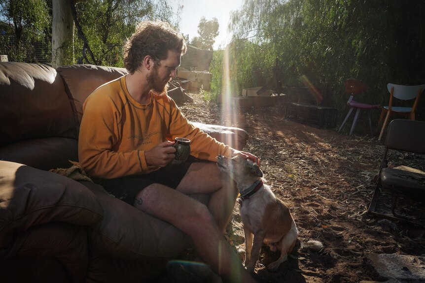 A man sitting in a lounge chair, outdoors, while holding a cup of tea and patting a dog. 