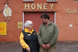 An older woman and a younger man stand outside a brick building smiling at eachother.