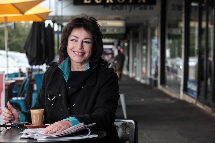 A woman with dark hair, wearing a teal shirt and black jacket, sitting at a table outside a cafe, with a coffee and newspaper.