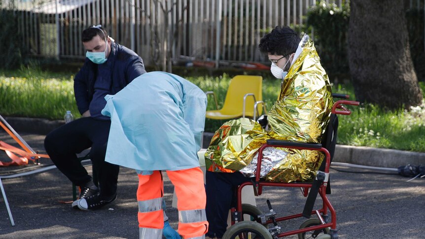 A health worker helps a man in a space blanket and face mask into a wheelchair