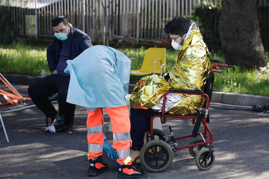 A health worker helps a man in a space blanket and face mask into a wheelchair