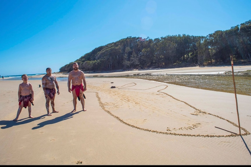 Three Indigenous men in loin cloths on beach inlet with giant whale drawn into sand, trees in back.