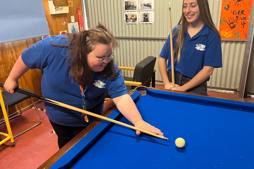 Two smiling young women in blue t-shirt, black pants playing pool, an orange poster behind them.
