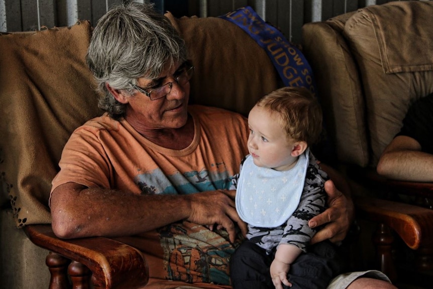 Garry Moratti sits on a chair holding his baby grandson