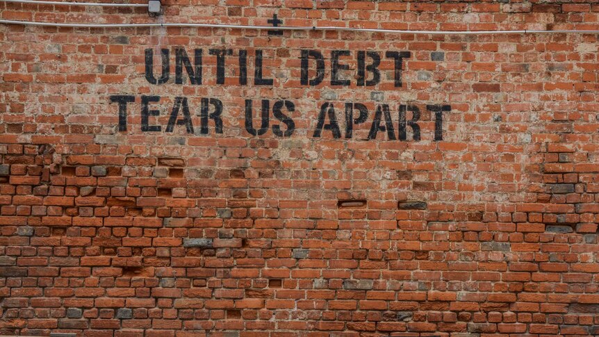 A brick wall with black writing spray-painted on it, saying 'until debt tear us apart'.