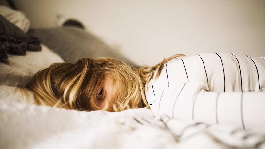 Woman lying face down on bed with her hair obstructing her face and looking tired to depict sleep problems.