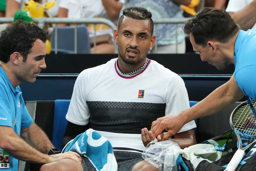 Nick Kyrgios receives treatment from trainers while sitting down on the side of the court.