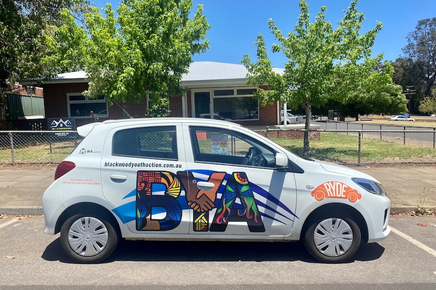 A small white car with a large BYA logo parked out the front of a brick building. 