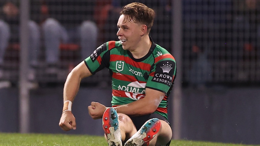 A South Sydney NRL player sits on the ground ans gestures as he celebrates scoring a try.