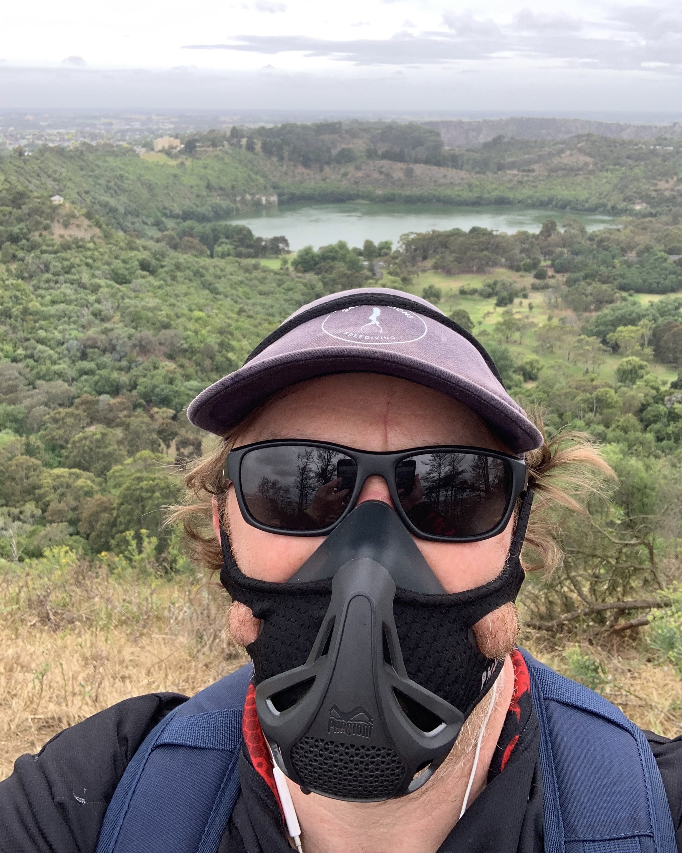 A man takes a selfie on top of a hill wearing a hat, glasses and a black mask.