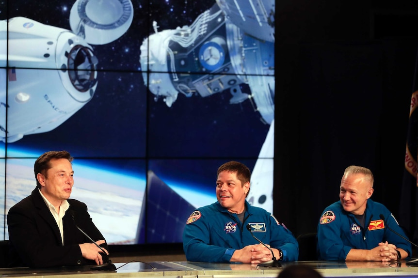 Elon Musk and two NASA officials sit at a table in front of a picture of the Dragon capsule