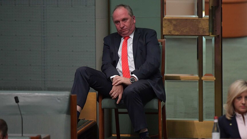 barnaby joyce sitting in a chair slouched in parliament wearing a black suit with a red tie his face is red