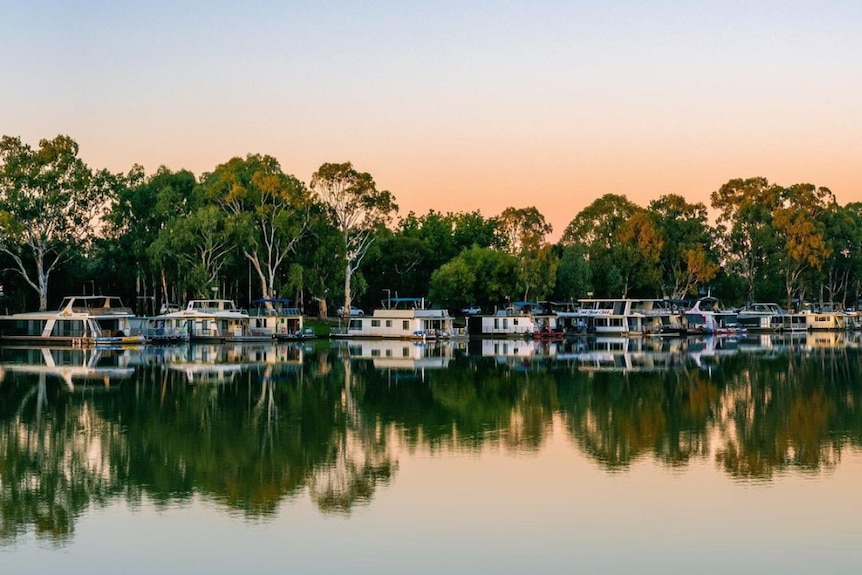 A river reflection with houseboats and tall trees lined along the blue and orange skyline 