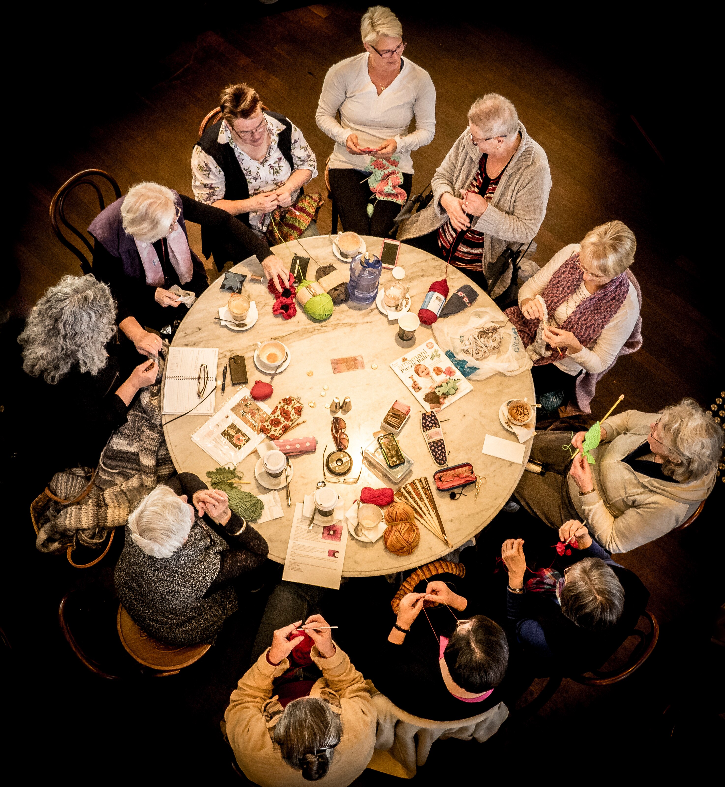 A group of women from above, sitting around a round table with cups of tea and knitting projects.