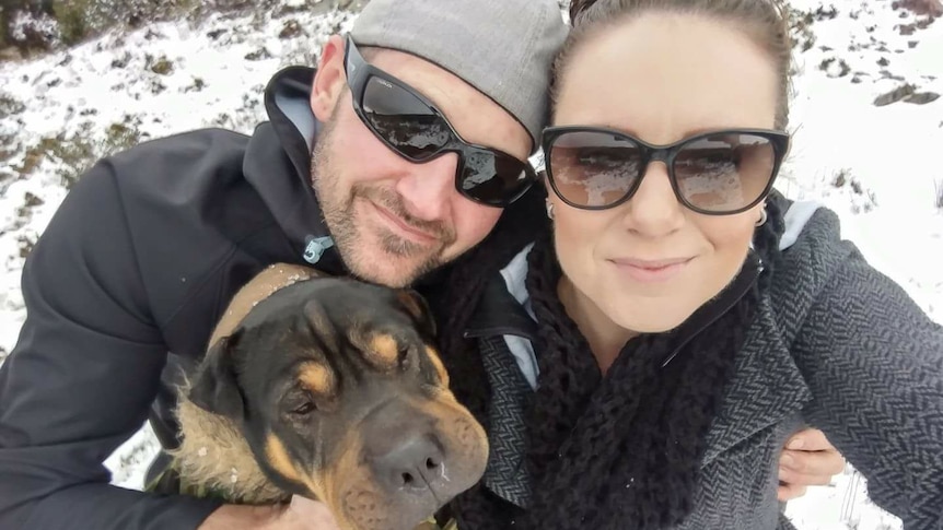 A man, woman and dog stand in the snow and smile at the camera.