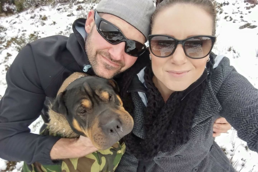 A man, woman and dog stand in the snow and smile at the camera.