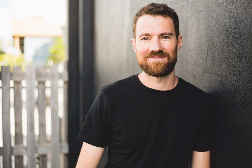 Man with brown hair and eyes smiles at camera while leaning against a wall