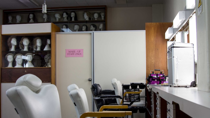 A room with chairs, lit mirrors and cabinets displaying mannequin heads wearing wigs.