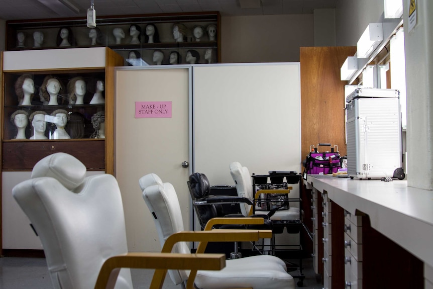 A room with chairs, lit mirrors and cabinets displaying mannequin heads wearing wigs.