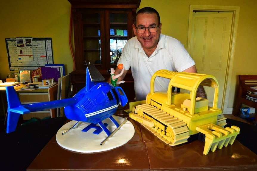 Middle-aged man poses with a digger and helicopter cake.