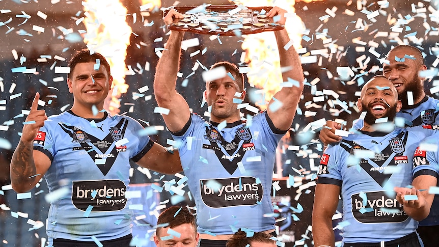 James Tedesco lifts the State of Origin shield as confetti falls. Latrell Mitchell and Josh Addo-Carr are next to him.
