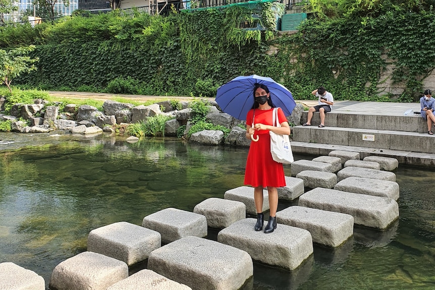 Sneha Karri stands on a stepping stone on a pond wearing a mask and holding an umbrella in front of a garden.