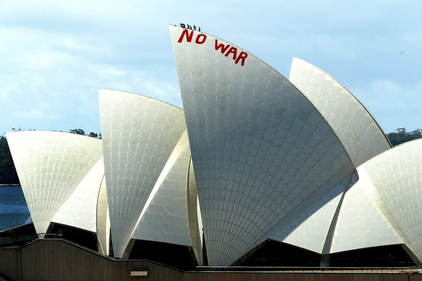 Wide shot of the six people at the top sail of the Sydney Opera House. A "No War" slogan is painted is painted in red in caps.