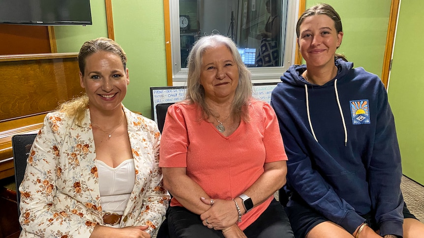 Three women sit side by side in a radio studio smiling at the camera.