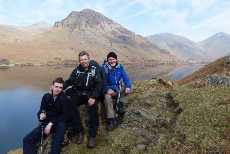 On a clear day, three men in hiking gear sit on a craggy rock in front of a mountains and a lake.