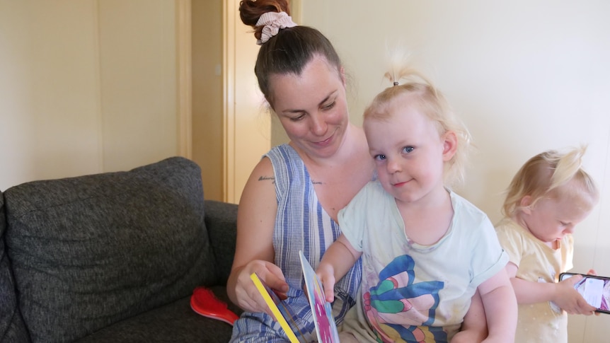 A mother reads a book to her preschool daughter while her twin sister plans on a phone