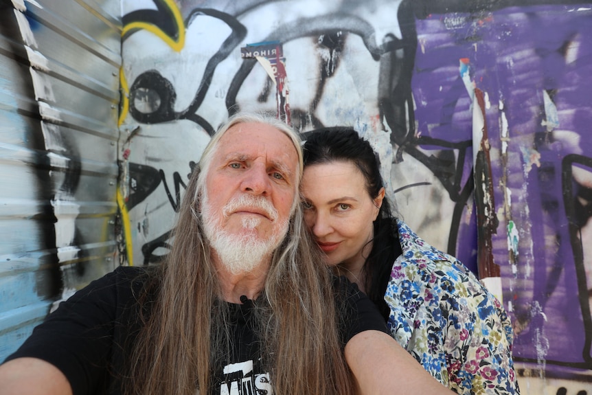 A man with long white hair and a grey beard takes a selfie with a woman with brown hair.