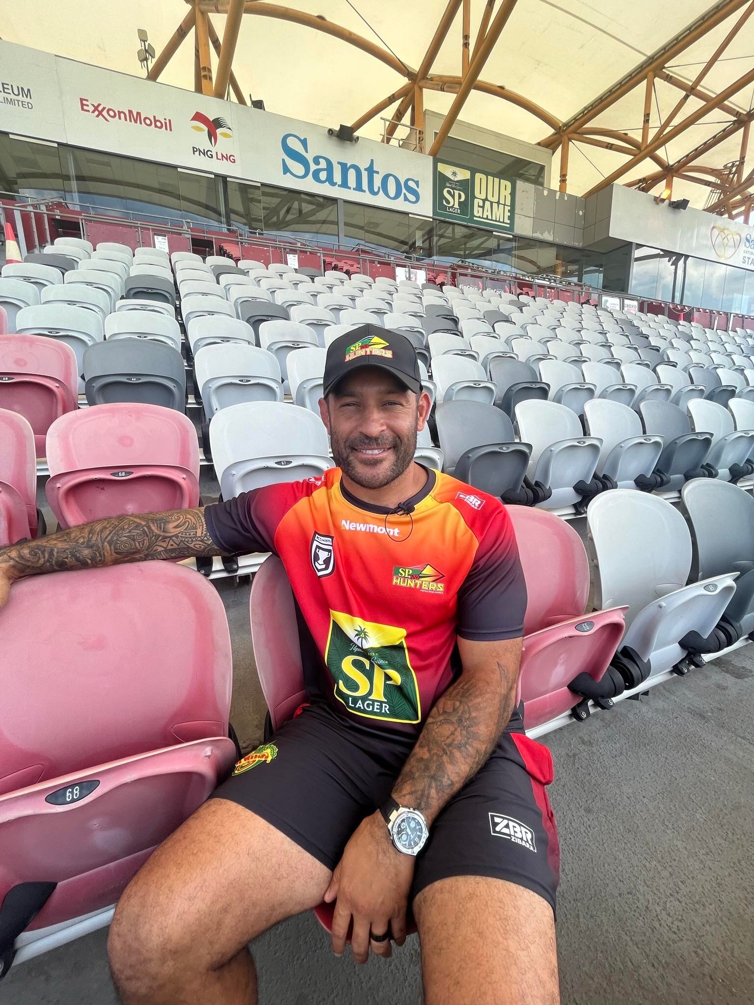Man wearing PNG Hunter shirt and shorts sits on stadium seats at a Papua New Guinea stadium smiling at the camera