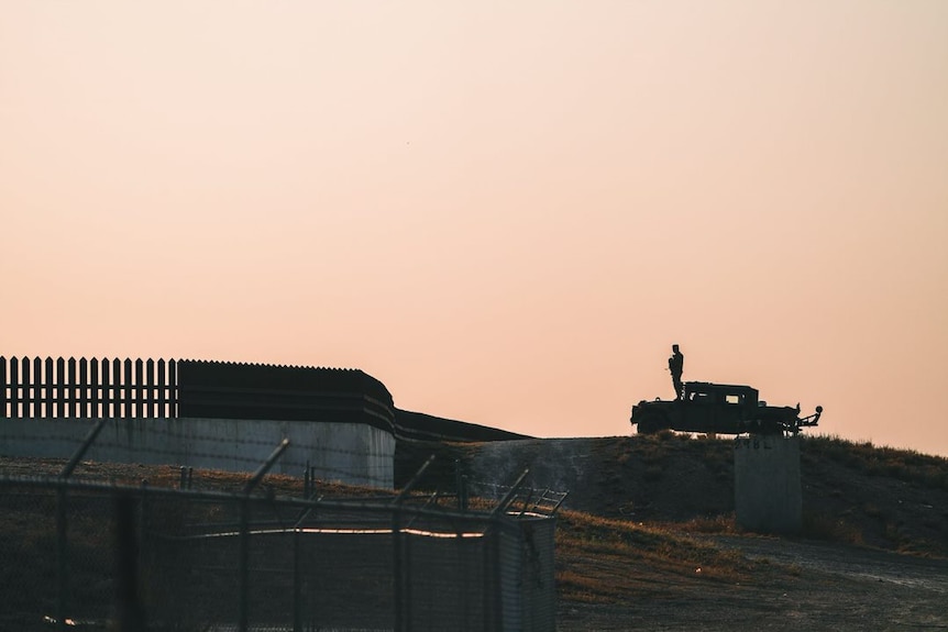 A person in shadow stands on a car as he looks over a stretch of wall.