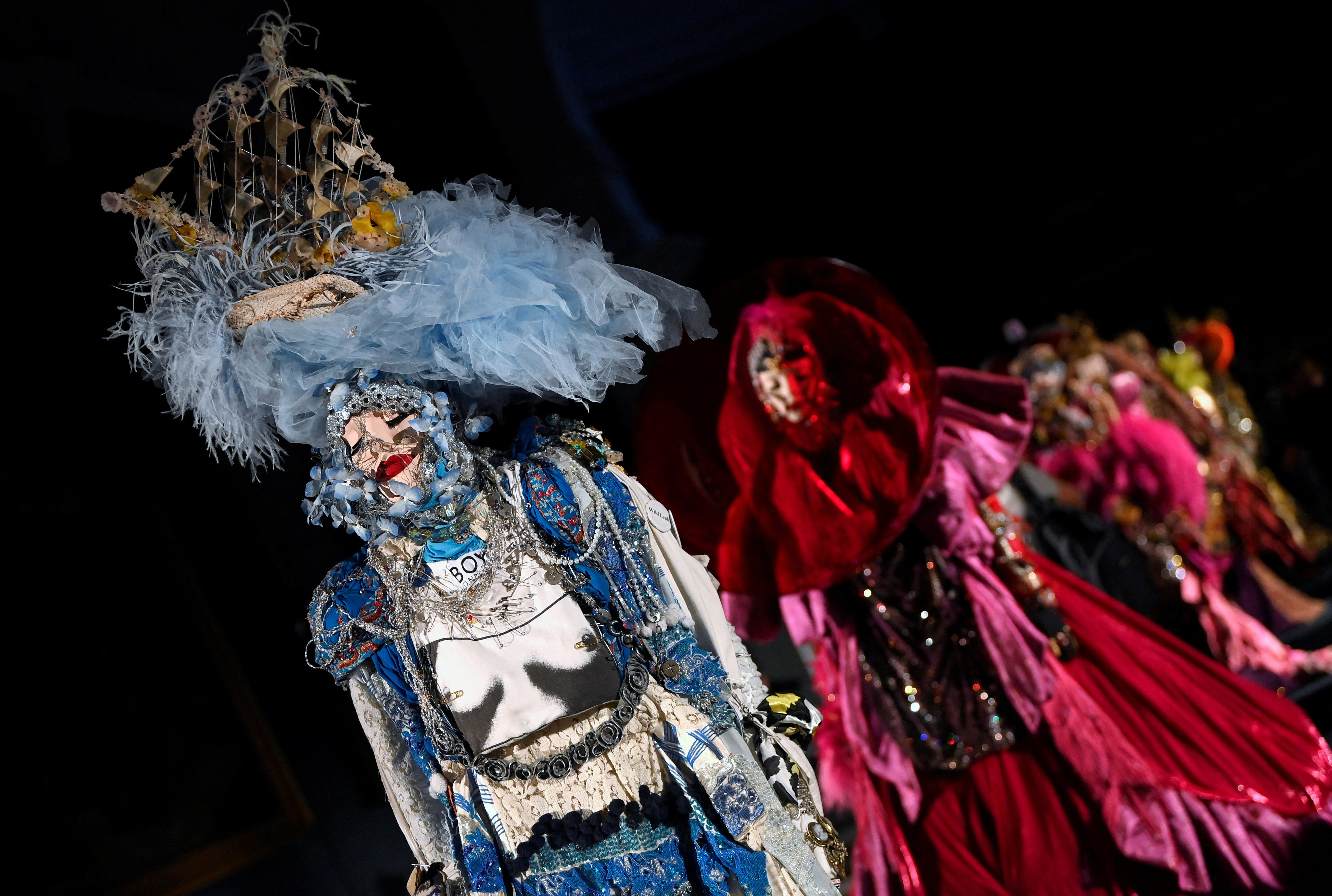 Mannequins wearing colorful and extravagant yet haunting costumes are lined up facing the camera.