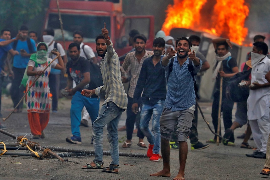 A group of men riot in front of a burning vehicle.