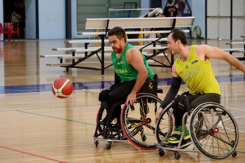 Two young men wearing green and gold uniforms play wheelchair basketball.