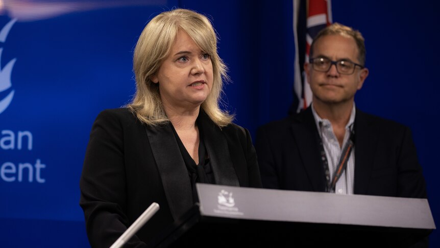 Blonde woman standing at a lectern speaking with a white man in glasses behind her. 